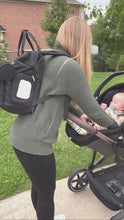Load and play video in Gallery viewer, Video showing a women using a diaper bag with her baby. She opens the changing stations and places the baby on it for a nap while sitting on the grass. After she packs the diaper bag and attaches is to the stroller using the stroller straps.
